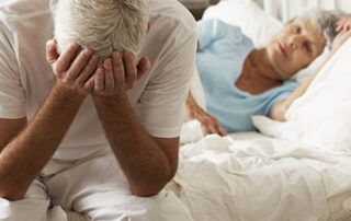 Worried Senior Man Sits On Bed Whilst Wife Sleeps