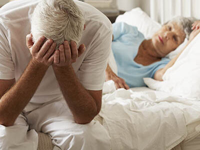 Worried Senior Man Sits On Bed Whilst Wife Sleeps