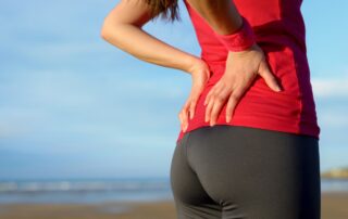 A lady with a painful back standing on the beach, clutching her lower back with her hand | Denver Stem Cell